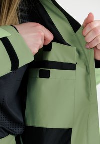 Vaaleanvihreä vedenpitävä takki, jossa on musta vuori. Takissa on kaksi ulkoista taskua, joissa on mustat tarranauha suljettavat läpät, ja sen pinta on sileä.