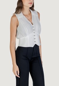 Blusa bianca senza maniche con pieghe frontali, colletto e sei bottoni lucidi. Indossata con pantaloni neri a vita alta, sartoriali.