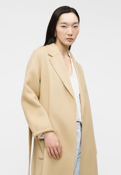 Γυναίκα με μακριά σκούρα μαλλιά φοράει μπεζ μακρύ παλτό, λευκό πουκάμισο, ανοιχτό μπλε παντελόνι και χρυσούς κρίκους σκουλαρίκια σε απλό λευκό φόντο.