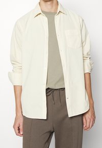 Camicia di corduroy beige chiaro con bottoni frontali, taschino sul petto e maniche arrotolate, abbinata a una maglietta grigia e pantaloni marroni.
