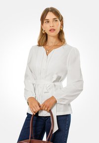 Blusa blanca de botonadura con cintura ajustable, mangas largas abullonadas y detalles de encaje en el frente. Combinada con jeans de mezclilla oscuros y un bolso marrón.