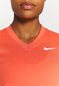 Nahaufnahme des unteren Gesichts und Halses einer Person, die ein korallenfarbenes V-Ausschnitt-Nike-Shirt mit einem weißen Swoosh-Logo auf der rechten Seite trägt.