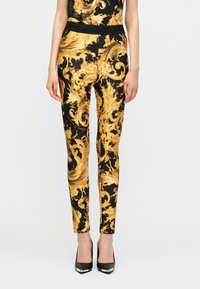 Γυναίκα με στενά παντελόνια σε μαύρο και χρυσό με μπαρόκ μοτίβο και ταιριαστό τοπ, συνδυασμένα με μαύρες μυτερές ψηλοτάκουνες γόβες.