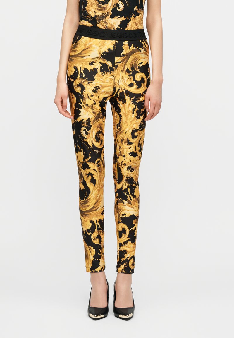 Γυναίκα με στενά παντελόνια σε μαύρο και χρυσό με μπαρόκ μοτίβο και ταιριαστό τοπ, συνδυασμένα με μαύρες μυτερές ψηλοτάκουνες γόβες.