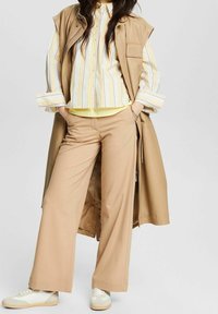 Beige ärmlös trenchcoat över en flerfärgad randig skjorta, krämfärgade byxor och ljusa sneakers. Nyckelfunktioner inkluderar vida ben och lager på lager-texturer.