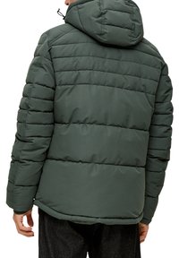 Man in een donkergroene gewatteerde jas met capuchon, van achteren gezien, staande tegen een witte achtergrond.