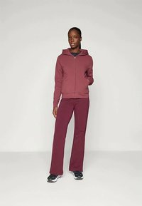 Bordo zip-up kapucar s kapuco, v kombinaciji z usklajenimi hlačami. Tkanina se zdi mehka z gladko teksturo. Teniske dopolnjujejo videz.