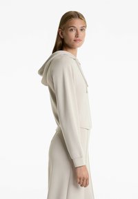 Hanorac scurt de culoare bej deschis, cu glugă reglabilă, mâneci lungi și textură fină. Prezintă un design elegant și cusături minime.