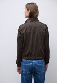 Femme aux cheveux bruns raides portant une veste en daim marron foncé et un jean en denim bleu, se tenant de face devant un mur blanc uni.