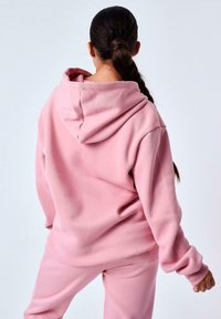 Sudadera de forro polar rosa con capucha ajustable, que presenta un corte relajado y mangas largas, combinada con pantalones de chándal rosa a juego. Textura suave.