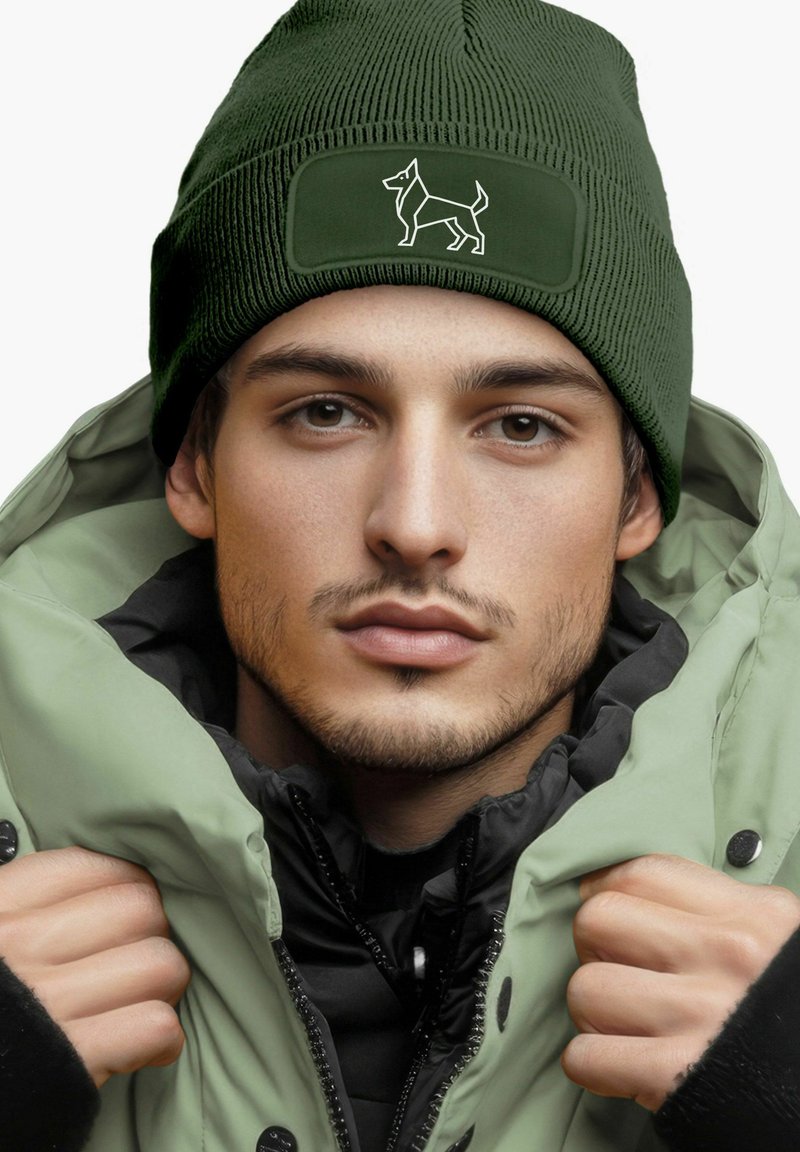 Dark green ribbed knit beanie with a rectangular patch featuring a white dog silhouette; model wearing a light green puffer jacket.