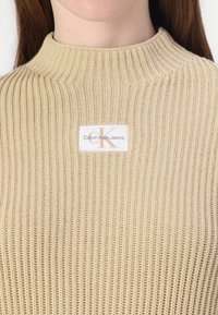 Suéter de punto acanalado beige con cuello alto. Presenta una etiqueta blanca con el logotipo de "Calvin Klein Jeans" en tonos beige y negro.
