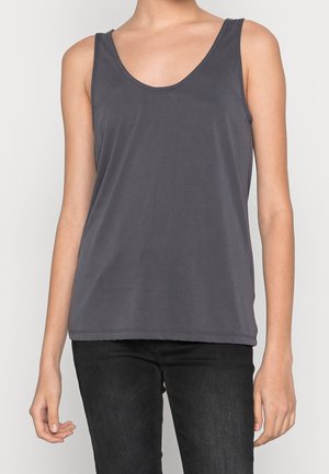 Kobieta w ciemnoszarej bezrękawnikowej koszulce z okrągłym dekoltem i czarnych spodniach, stojąca na jednolitym jasnym tle.