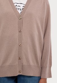 Beige cardigan with a V-neck, six brown buttons, and ribbed cuffs. Fabric appears soft with a smooth texture. Worn over a white graphic shirt.