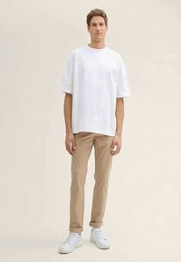 T-shirt branco de manga curta, corte relaxado, usado com calças bege. Modelo em pé, com ténis brancos, contra um fundo neutro.