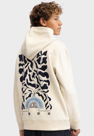 Persona joven con pelo rizado que lleva una sudadera con capucha color crema con un diseño abstracto de planta en negro y el texto "Grow into the wild" en la espalda.