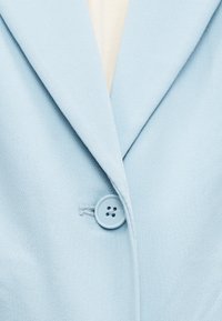 Ανοιχτό γαλάζιο σακάκι με ένα κουμπί, με φαρδιές πέτρες, που αφήνει να φαίνεται ένα λευκό πουκάμισο από μέσα.