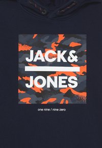 Námorně modrá mikina s kapucí s obdélníkovou grafikou v maskáčovém vzoru v šedé, černé a oranžové barvě. Text "JACK & JONES" je vyveden výrazně.