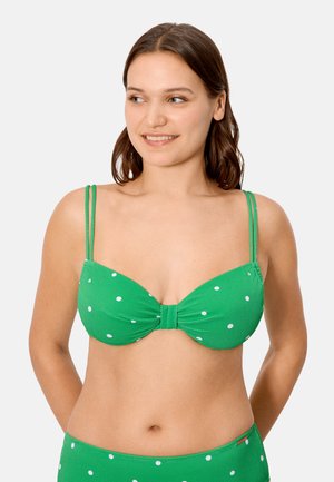 Mujer con un bikini verde con lunares y doble tirante, mirando ligeramente hacia su izquierda contra un fondo liso y claro.