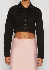 Woman wearing a black cropped button-up jacket with two chest pockets and a high-waisted, light pink skirt showing midriff.