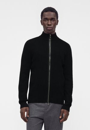 Homme portant un pull zippé noir côtelé et un pantalon gris, debout devant un fond clair uni, face à l'avant.