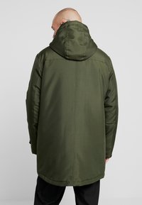 Oliwkowa parka z kapturem, o gładkiej teksturze, długości do pełnej długości i subtelnych detalach przeszyć. Zaprojektowana dla zapewnienia ciepła i ochrony.
