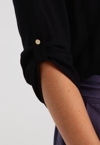 Manga de camisa negra con puño enrollado, que presenta un botón dorado redondo. Textura suave, diseño ajustado y caída sutil.