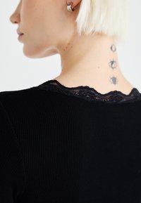 Top preto com textura canelada, apresentando uma delicada renda preta na gola. O design do tatuagem visível consiste em formas geométricas ao longo do pescoço.