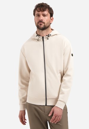 Man draagt een beige hoodie met ritssluiting en trekkoorden en bruine broek, staand tegen een effen witte achtergrond.