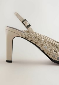 Sandale à talons hauts beige avec des brides tressées et une boucle réglable à la cheville, dos fermé et bord de semelle noir.