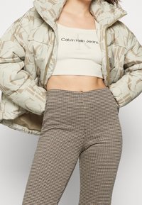 Puffjacka i ljusbeige med brun abstrakt mönster, kombinerad med en krämfärgad croptop och bruna rutiga högmidjade byxor.