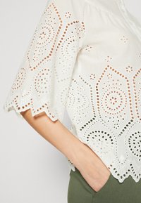 Blouse blanche pour femme en dentelle eyelet avec ourlet et manches festonnés, présentant des motifs découpés géométriques et floraux.