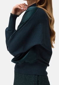 Jersey de color verde oscuro con cuerpo de punto texturizado, acentos de satén suave, mangas anchas y bajo de canalé.