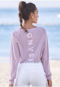 Helles rosa Langarmoberteil mit lockerer Passform und vertikalem weißen "SAND"-Druck. Kombiniert mit weißen Leggings, die einen glatten Bund haben.