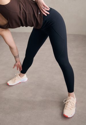 Schwarze Sportleggings mit glatter Textur und enganliegendem Design, kombiniert mit hellbeigen Sneakers mit rosa Akzenten und Schnürsenkeln.