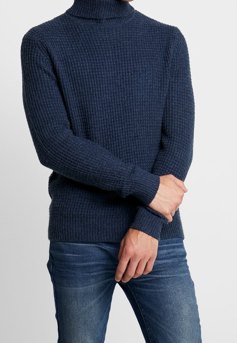 Marinblå stickad polotröja med strukturerat mönster och ribbade muddar, kombinerad med ljusa jeans i denim.