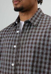 Bruin-grijs geruite flanellen blouse met een knoopsluiting aan de kraag, voorzien van witte knopen en een zachte textuur. Casual ontwerp met zakken op de borst.