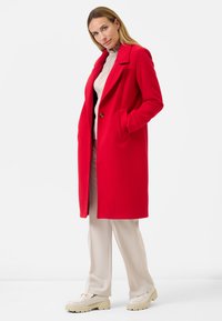 Rode wolmix jas met een geknipt kraag, zijzakken en een enkele knoopsluiting, gecombineerd met beige broeken en crèmekleurige schoenen.