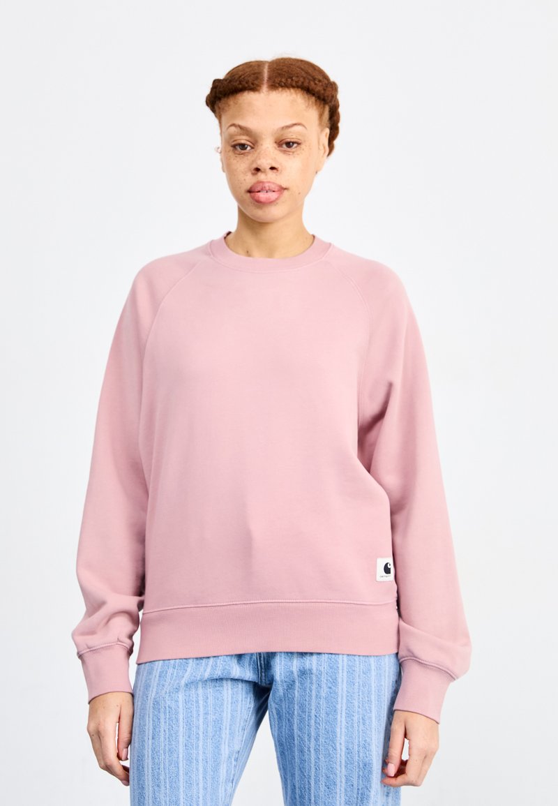 Sweat-shirt rose clair avec un col rond côtelé et de longues manches raglan. Comprend une petite étiquette de logo au bas de l'ourlet. Associé à un pantalon bleu rayé.