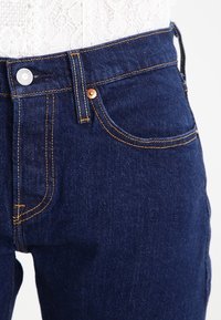 Jeans in denim lavaggio scuro con hardware in tonalità rame, dettagli di cuciture a vista e vestibilità slim. Presenta tasche anteriori e chiusura con bottone.