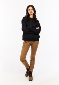 Zwarte rits-hooded jacket, bruine gevormde broek en tan kleurige wandelschoenen. De jacket heeft een gladde textuur; de broek is lichtgewicht.