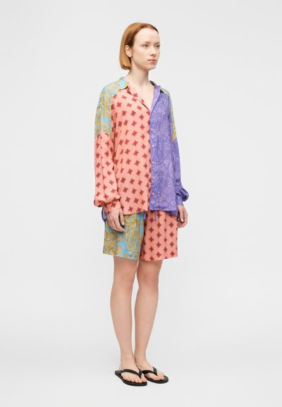 Γυναίκα που στέκεται, φοράει πολύχρωμο πουκάμισο με μακριά μανίκια σε σχέδιο παζλ και αντίστοιχα σορτς, μαζί με μαύρα σανδάλια, σε απλό φόντο.