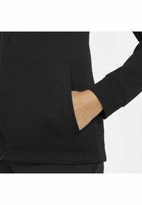 Hand som bär ett svart långärmat plagg med handen delvis i den främre fickan mot en vit bakgrund.