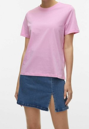 Femme portant un t-shirt rose uni à manches courtes et une mini-jupe en jean bleue avec une fente à l'avant, les mains détendues le long du corps.