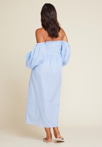 Blå og hvit gingham avkuttet skulderkjole med pufferm, smocket midje og lang, rettskåret skjørt. Flate sandaler synlige.