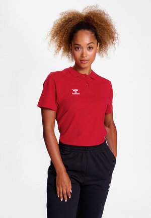 Rød polo-shirt med krave, der har et hvidt logo på venstre bryst. Kombineret med sorte bukser med elastisk talje. Glat bomuldsstruktur.