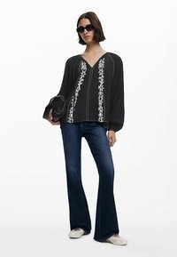 Sort bluse med hvid blomsterbroderi, der løber lodret, med lange ærmer, kombineret med mørke denimjeans med vidde og slip-on sko.