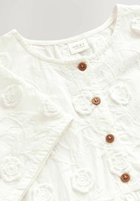 Witte top met geborduurde bloemen, voorzien van een ronde halslijn, korte mouwen en bruine knoopsluitingen aan de voorkant. Zachte textuur.