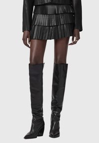 Μίνι μαύρη δερμάτινη φούστα με πλισέ υφή, σε συνδυασμό με μαύρες δερμάτινες μπότες πάνω από το γόνατο, με μυτερή μύτη και χοντρούς τακούνια.