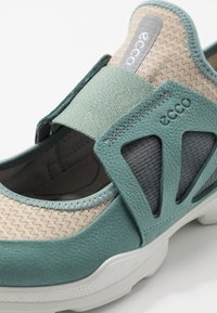 Turquoise and beige slip-on shoe with textile and synthetic upper, elastic strap, mesh accents, and a white durable rubber sole.
